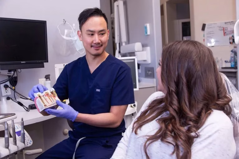 Dentist in blue scrubs discussing dental implant options with a patient in a dental office, showcasing a model of dental implants.