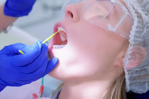 Child receiving fluoride treatment at dental office, dental professional applying fluoride with applicator, protective eyewear and dental cap visible.