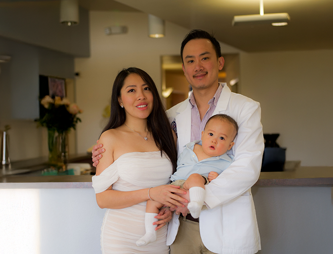 Family portrait featuring a woman in a white dress, a man in a white blazer and purple shirt, holding a baby in a light blue outfit, set in a welcoming dental office environment.